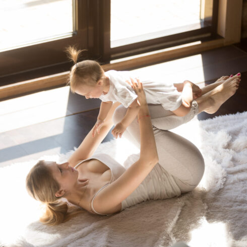 Eine Mutter die mit ihrem Baby Yogaübungen auf einer Decke auf dem Boden macht.